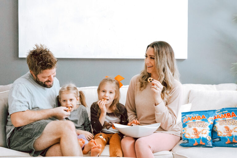 A family of four sits together on a light-colored couch, smiling and eating peanut butter snacks from a bowl. A bag of BAMBA Peanut Butter Puffs is visible beside them, suggesting a relaxed family snack time at home.