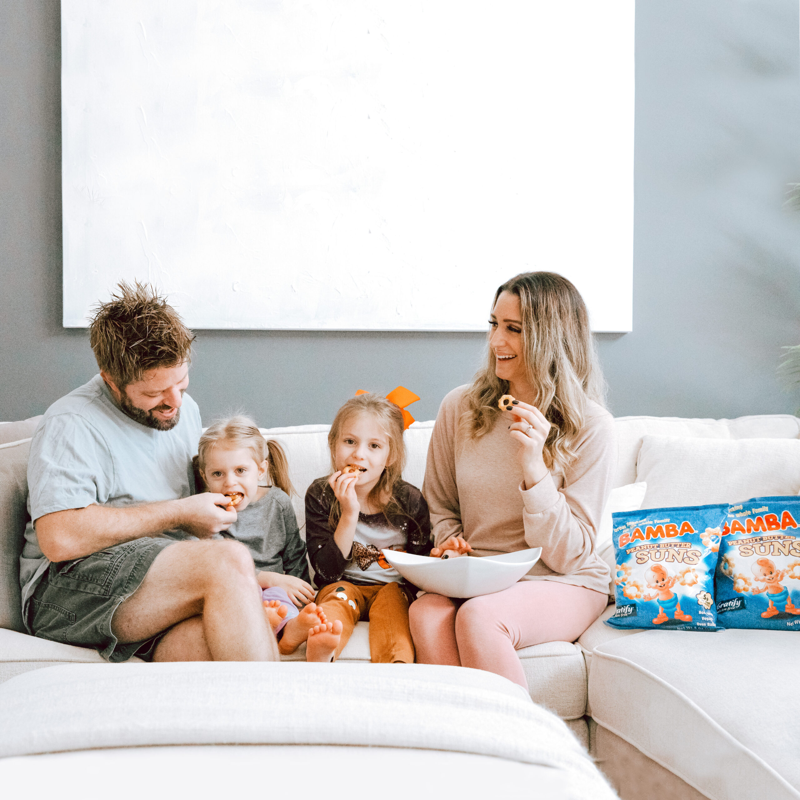 A family of four sits together on a light-colored couch, smiling and eating peanut butter snacks from a bowl. A bag of BAMBA Peanut Butter Puffs is visible beside them, suggesting a relaxed family snack time at home.