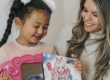 A mother and young daughter smile while looking at a colorful toy package featuring a fantasy-themed character. The child holds a pink toy case, and both appear excited as they examine the new toy together indoors.