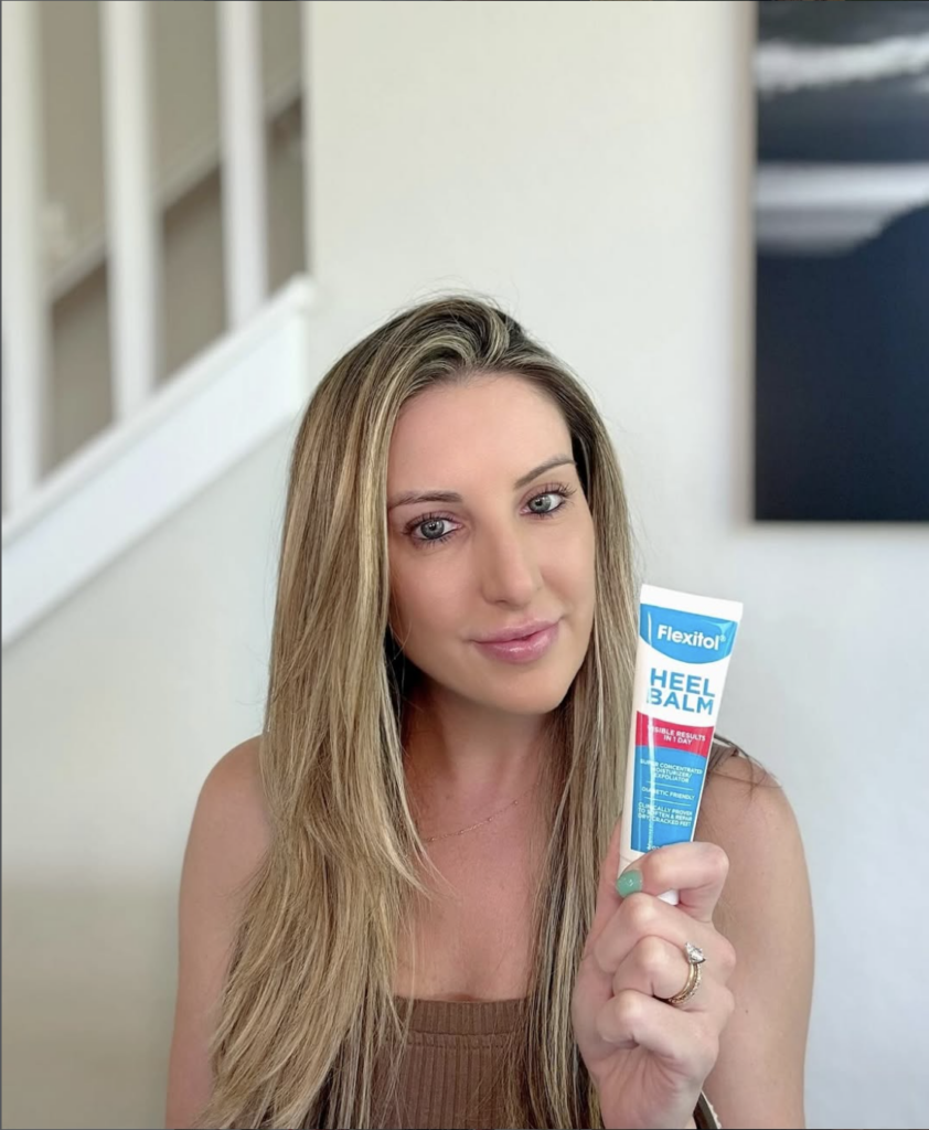 A woman indoors holds up a tube of Flexitol Heel Balm toward the camera, showcasing the product while looking directly at the viewer.